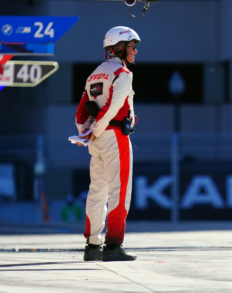 Race car driver in a white-and-red racing suit and helmet, standing on the pit lane while holding papers.