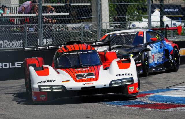 Porsche race car leading a red-and-white livery on a race track with a blue Corvette behind, barriers and spectators in the background
