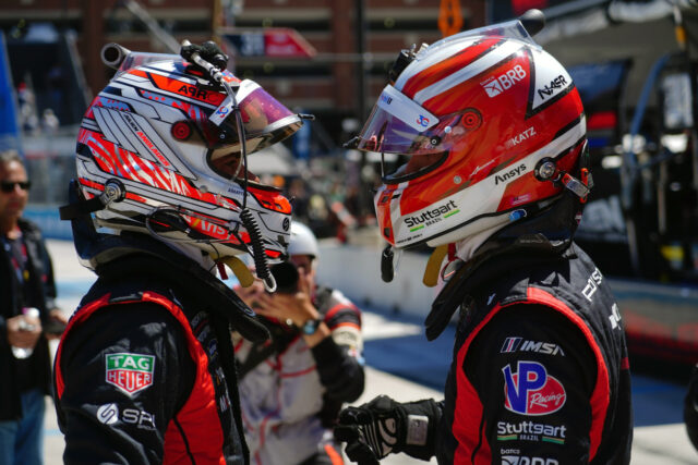 Two race car drivers in red-and-black suits shake hands in a pit lane, wearing colorful helmets with sponsor logos in the background.