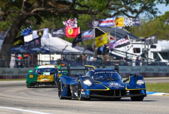 Blue Aston Martin race car leads on a sunlit track, a green-yellow car close behind with flags and tents in the background.