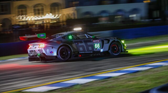 Nighttime Ford race car #66 speeding along a lit race track, green front lights and blurred background.
