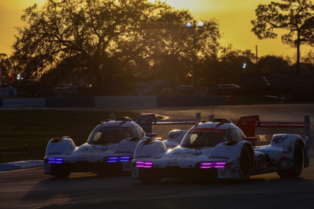 Race cars lined up at the track during sunset, with a yellow sky and silhouetted trees in the background, Porsche branding visible on the cars.
