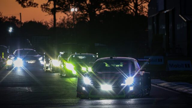 Line of cars at night with bright headlights on a wet road, orange sky in the background.