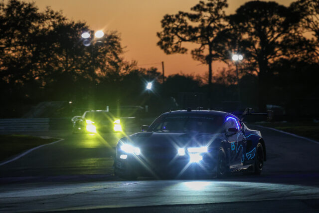 Race cars speeding on a track at dusk with bright headlights and orange sky behind silhouettes of trees. Option 1: A blue race car leads on a dim track with glare from its headlights and other cars in the background. Option 2: Night-time race scene with two cars illuminated by front headlights, smoke or glare, and dark treelines. Option 3: A blue race car number 65 driving at high speed on a curving track as stadium lights glare ahead. Option 4: Polished race car on a racing circuit at sunset, headlights blazing and spectators faintly visible in the background.