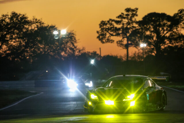 Two police cars with flashing yellow and blue lights in a dark parking lot, orange dusk sky and tree silhouettes in the background.
