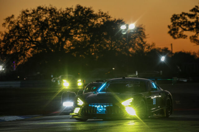Race car on a dimly lit track at dusk with bright headlights and orange sky in the background.