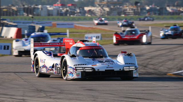 White Porsche race car with red accents leading a pack on a racetrack, sponsor logos visible.