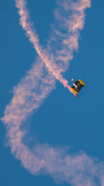Skydiver with a yellow and black parachute releasing a pink smoke trail against a clear blue sky