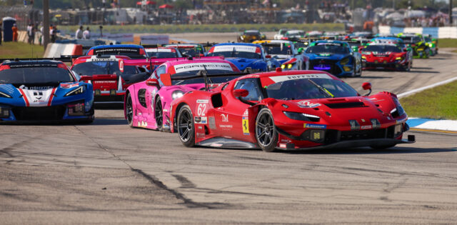 Group of colorful race cars cornering on a track with a red Ferrari in the foreground and a crowded pit area in the background.