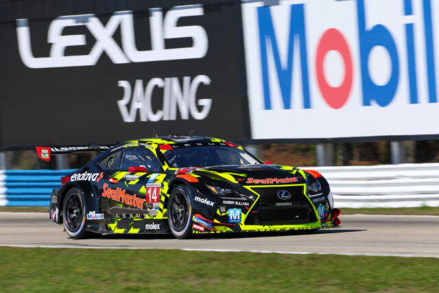 Racing car on a track with bright livery and sponsor decals speeding past blue barriers and a large advertisement behind.