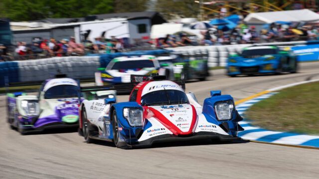 Race cars speeding around a track with the lead car in white, red, and blue lapping others as spectators watch from the pit wall in the background
