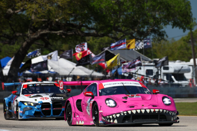 Pink Porsche race car with a shark-mouth design leads a pack on a track with blue and white cars behind, colorful flags in the background.