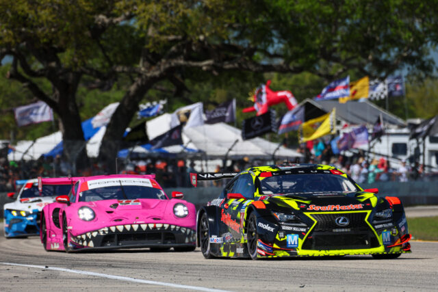 Pink Porsche with a shark-mouth livery leads a pack of race cars on a track, colorful flags overhead and a cheering crowd in the background.] , should be: