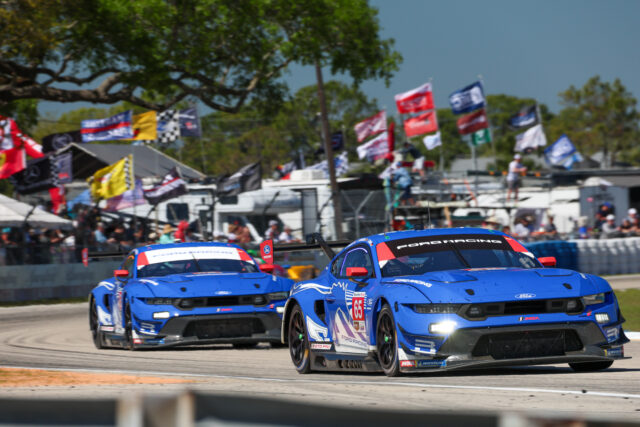 Two blue Ford Racing cars race side by side on a track, with a crowded pit area and colorful flags in the background.