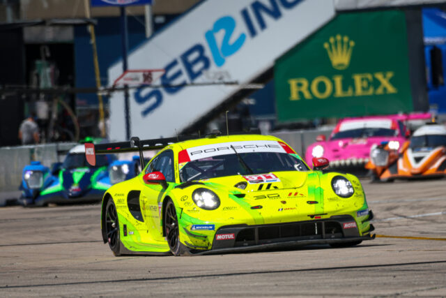 Bright yellow Porsche race car speeding on a track with a Rolex billboard in the background, pit lane scenes nearby.