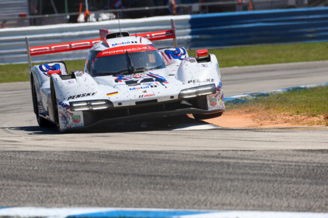 White Porsche race car with red and blue decals speeding along a race track, barriers in the background.