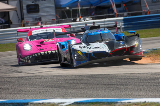 Two race carsโpink Porsche and blue BMWโbattle for position on a racetrack with blurred spectators in the background.