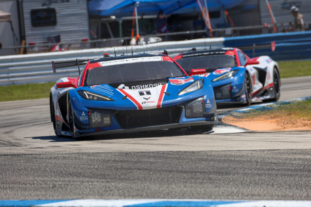 Two race cars, blue and red, speed around a curve on a race track with a pit building in the background.