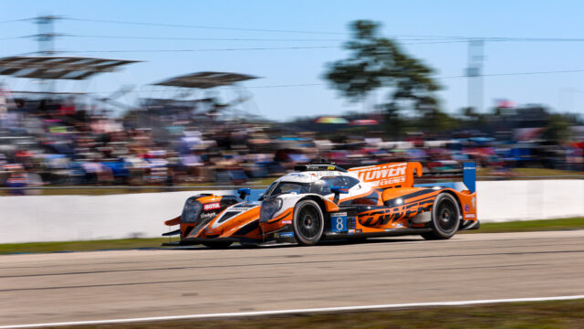 Orange and white race car sprinting along a racetrack with blurred grandstands in the background (car #8).