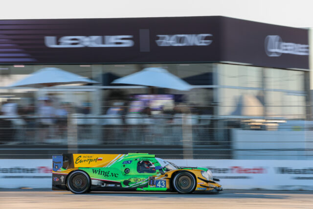 Green and yellow race car speeding on a track with blurred grandstands in the background, sponsor logos visible on the car.