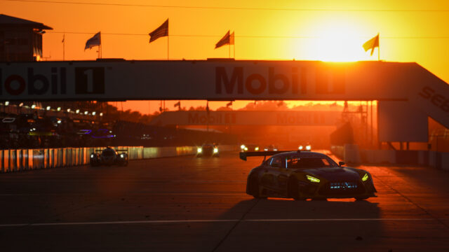 Race car on a track at sunset with an orange sky, Mobil 1 banners overhead and blurred spectators in the background.