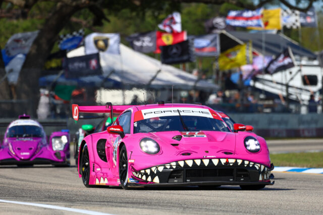 Pink Porsche race car with shark-teeth graphics leads a lineup on a race track, with tents and colorful flags in the background.