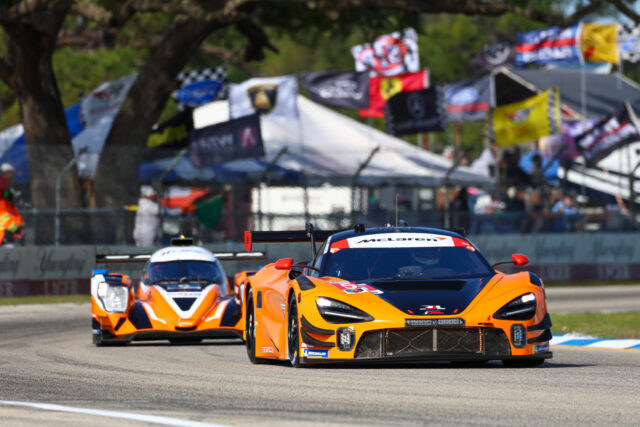 Orange McLaren race car leading a pack on a sunny circuit, with another orange car behind and blurred pit-area banners in the background.