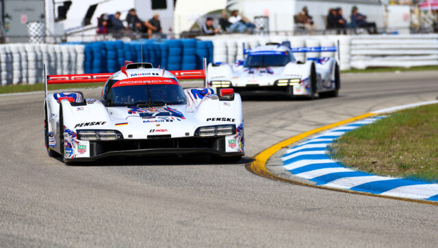 White Porsche race car leads a pack around a racing circuit, blue-and-white barriers and blurred spectators in the background.