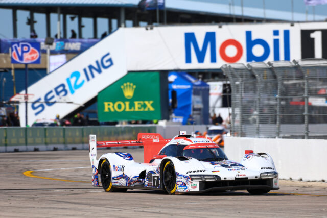 White Porsche race car driving on a track with sponsor banners and barriers in the background, blurred grandstand area visible behind.