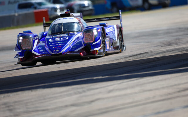 Blue prototype race car speeding around a track, sponsor logos visible on the chassis and wingtips, camera angle from the front-left.