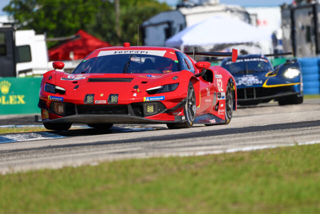 Front view of a red Ferrari race car speeding on a racetrack, with a blue car close behind in the background.