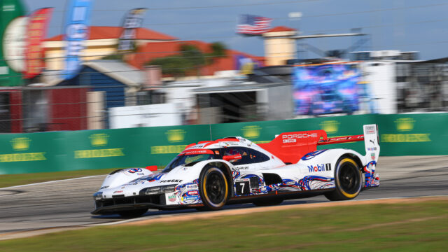 Two white Porsche race cars with red and blue livery sprint along a racetrack, sponsor decals visible, blurred background barriers and stands behind them.