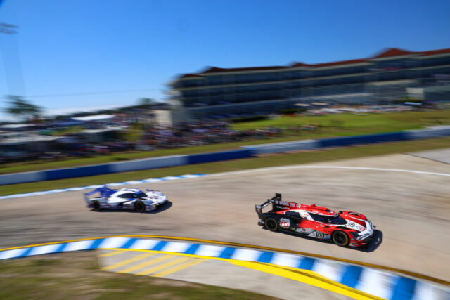 Two race cars speed around a curve on a sunny outdoor track, with a blurred grandstand and blue sky in the background.