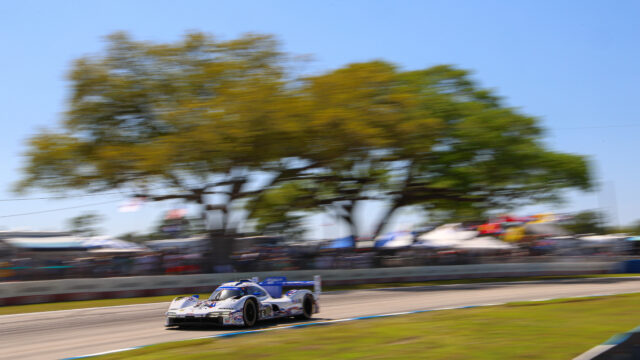 White and blue race car speeding on a track with blurred trees and spectators in the background.