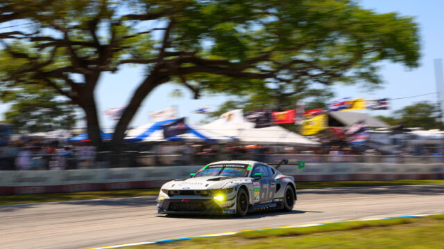 White race car number 66 with green accents speeding along a track, with blurred crowd and trees in the background due to motion blur.