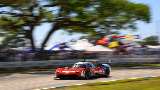 Red race car leading a blurred group on a racetrack amid trees and banners, motion blur conveying high speed.