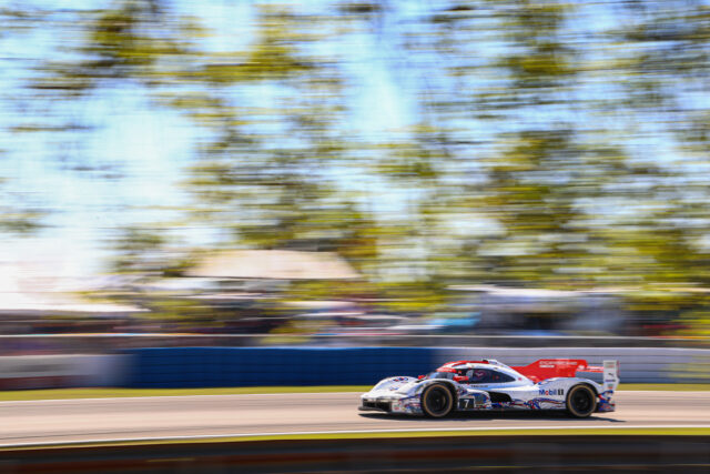 Two white Porsche race cars speeding along a race track with a blurred background of trees, Mobil 1 branding visible on the cars.