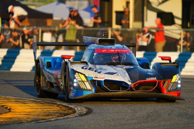 Blue and white prototype race car with red accents turning a corner on a racetrack, spectators in the background.