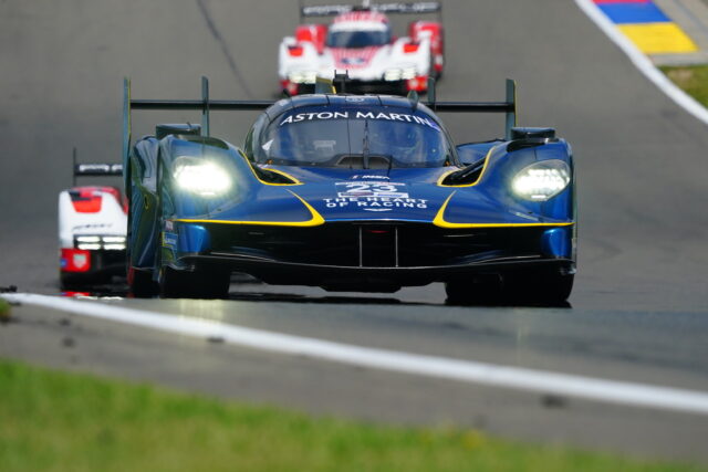 Blue Aston Martin racing car leading on a race track with other cars behind, headlights on, yellow accents on the body.