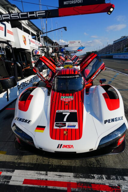 Red and white Porsche race car in the pit lane with gull-wing doors open, displaying sponsorship logos WeatherTech and TAG Heuer.