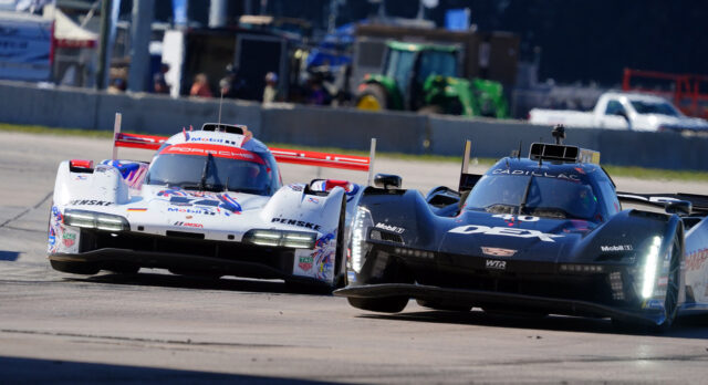 Two endurance race cars compete on a sunlit track, white Porsche ahead of a black Cadillac DPi-V.R, with pit buildings in the background.
