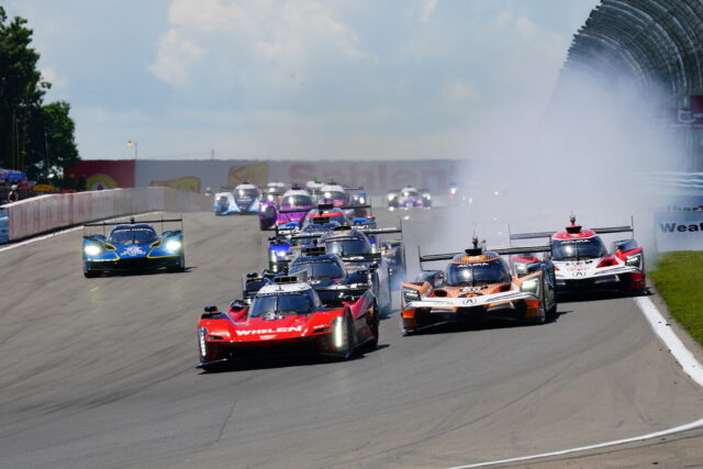 Red Whelen race car leads a pack of open-wheel race cars on a curving track, with tire smoke in the background and walls on the side.
