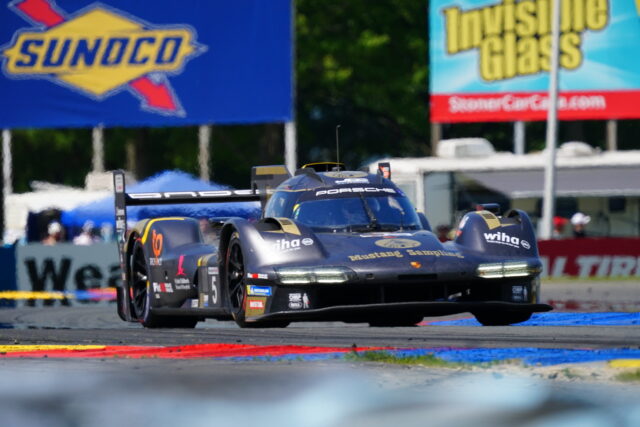 Porsche race car on a race track with dark blue and gold livery, sponsor decals, speeding past colorful track curbs, banners in the background.