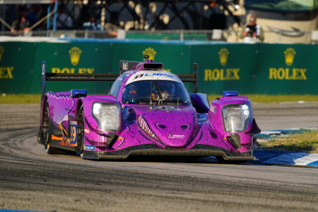 Purple Le Mans prototype race car (#19) corners a track with green Rolex banners in the background, smoke-free and focused on the apex.