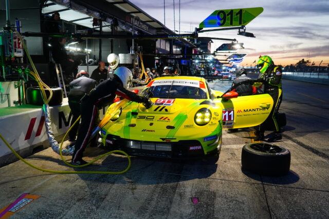 Pit crew refueling a bright yellow-green Porsche race car in the pit lane at sunset, with crew members and equipment nearby