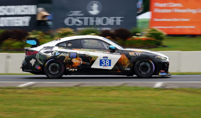 Race car number 38 speeding along the track with black-orange camouflage and sponsor decals on the side files.