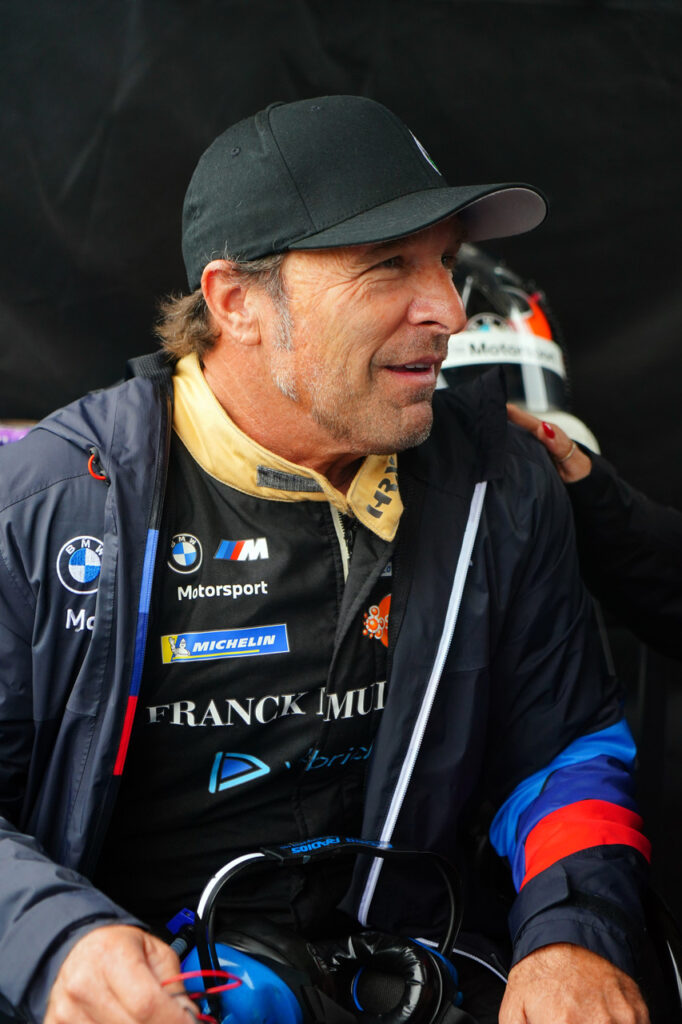 Older man in a racing jacket and cap, logos on his chest, seated with a helmet in the background.
