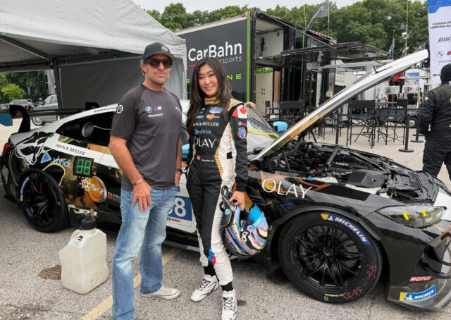 Man in a cap and sunglasses stands with a female race car driver in a white-black racing suit beside a race car with its hood open in the paddock areas behind tents and trailers.
