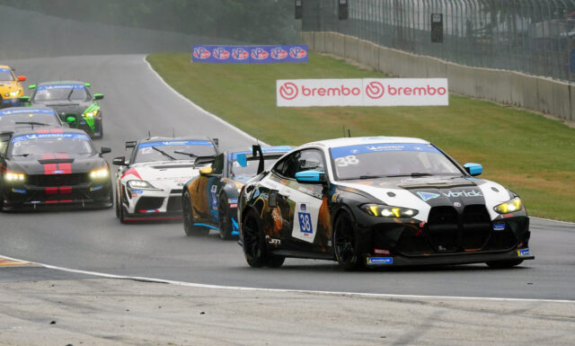 Race cars sprint around a wet track, led by a black-and-white car #38 with blue mirrors and sponsor decals on the right, as others follow closely in colorful liveries with a Brembo sign in the background.
